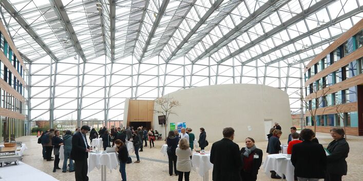 Veranstaltungsambiente im Forum des Umweltbundesamtes unter dem großzügigen Glasdach mit Blick nach draussen durch die Glasfront. Klar zu sehen sind die Teilnehmenden im Vordergrund, die sich während der Mittagspause unterhalten.