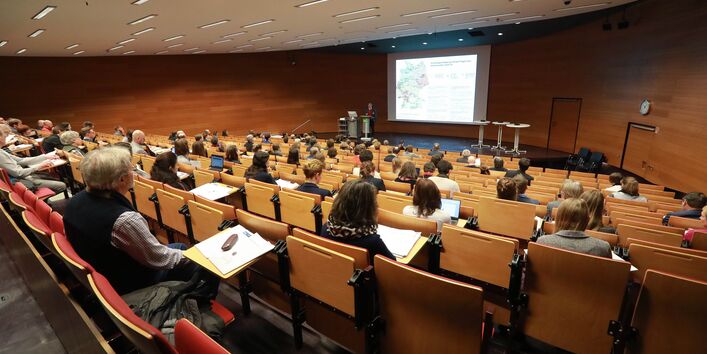 Veranstaltungsambiente im Hörsaal des Umweltbundesamtes. Der Blick geht nach vorn Richtung Podium zur Projektionsfläche. Ein Teilnehmener steht auf dem Podium am Stehpult und präsentiert seinen Vortrag. Die Teilnehmenden sitzen davor und hören zu. Der Hörsaal ist in warmen Holzfarben gehalten.
