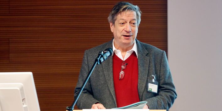 Ein Vortragender steht am Stehpult und spricht ins Mikrofon. Die Vortragsunterlagen hält er in der Hand. Der Vortragende trägt einen roten Pullover mit Jacket darüber.