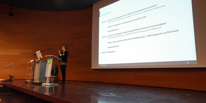 Vortragende steht auf dem Podium an einem Rednerpult und trägt vor mit einem Mikrofon in der Hand. Im Hintergrund ist das Veranstaltungsprogramm auf der Projektionsfläche zu sehen.