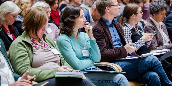Teilnehmende sitzen auf Stühlen in Reihen und beschäftigen sich mit Schriftgut in ihren Händen.