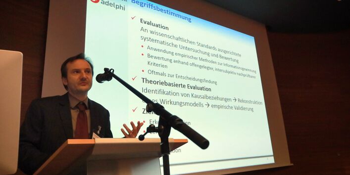 Vortragender trägt mit seinen Händen gestikulierend vor. Er steht am Rednerpult vor einem Mikrofon. Im Hintergrund ist seine Präsentation zu sehen zur Überschrift Begriffsbestimmung.