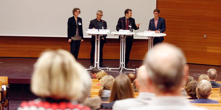 Teilnehmende diskutieren miteinander auf dem Podium. Sie stehen nebeneinander an Stehtischen aufgereiht. Der blick geht von hinten zwischen den Zuhörenden durch nach vorne.