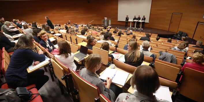 Veranstaltungsambiente im Hörsaal des Umweltbundesamtes: Der Blick geht nach vorne über die sitzenden Teilnehmenden hinweg zum Podium hin. Auf dem Podium stehen Diskutanten und Diskutantinnen an Stehtischen.