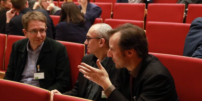 Teilnehmende sitzen auf den roten Sitzen im Hörsaal und sprechen miteinander. Ein Teilnehmer hebt dabei gestikulierend seine linke Hand.