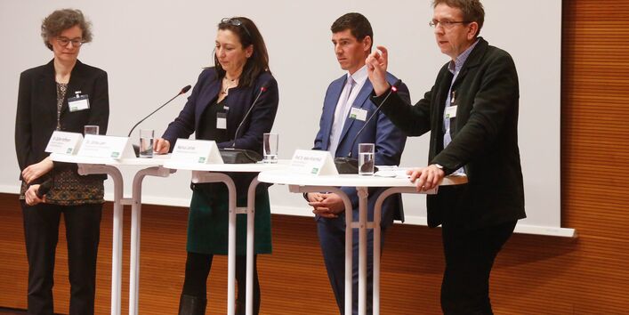 Teilnehmende stehen an Stehtischen auf dem Podium. Auf den Stehtischen stehen Gläser mit Wasser. Namensschilder und Tischmikrofone. Ein Teilnehmer spricht gestikulierend ins Publikum.
