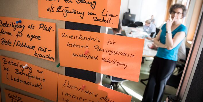 Im Vordergrund in groß sieht man zum Thema Beteiligung beschriftete, orangene Moderationskarten. Im Hintergrund steht eine Teilnehmerin im Workshopgruppenarbeitsraum vor den Arbeitstisch. Ein Teilnehmer sitzt schreibend am Tisch.