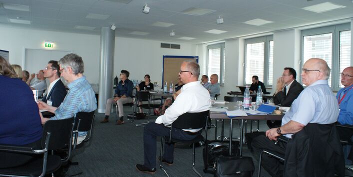 Teilnehmer sitzen im Arbeitsgruppenraum an Arbeitsgruppentischen mit dem Redner zugewandten Gesichtern.