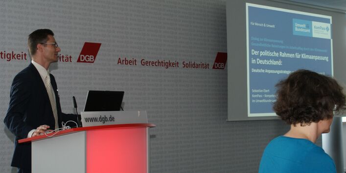 Vortragender steht am Rederpult und präsentiert im Hintergrund seitlich den Vortrag: Der politische Rahmen für Klimaanpassung in Deutschland.