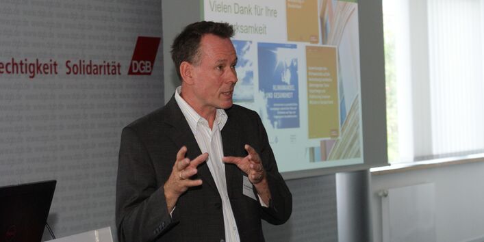 Vortragender steht am Rederpult und diskutiert gestikulierend mit den anderen Teilnehmern. Im Hintergrund ist der Abspann seiner Präsentation: Gesundheitliche Auswirkungen des Klimawandels in Deutschland