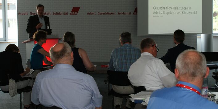 Vortragender steht am Rederpult und diskutiert gestikulierend mit den anderen Teilnehmern. Im Hintergrund ist das Startbild seiner Präsentation zu sehen.