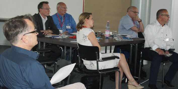 Teilnehmer sitzen im Arbeitsgruppenraum an Arbeitsgruppentischen mit dem Redner zugewandten Gesichtern.