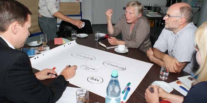 Teilnehmer sind am Gruppenarbeitstisch und diskutieren miteinander. Ein Teilnehmer schreibt Diskussionsergebnisse auf Posterpapier, welches auf dem Tisch liegt.