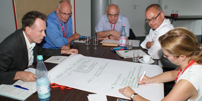 Teilnehmer sitzen im Arbeitsgruppenraum am Arbeitsgruppentisch und schauen auf das, was eine Teilnehmerin auf dem auf dem Tisch liegendes große Blatt Papier schreibt.