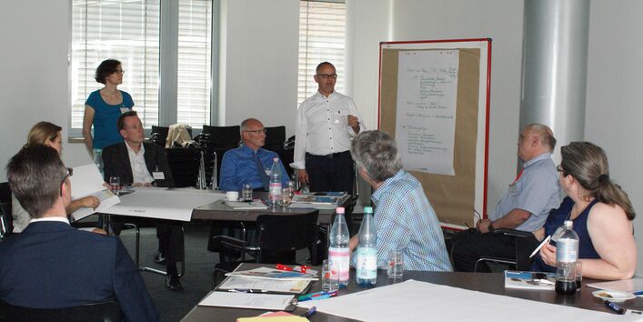 Vortragender steht an einer Moderationswand und trägt vor. Teilnehmer sitzen an den Gruppenarbeitstischen davor und hören aufmerksam zu.