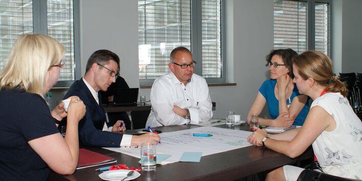 Teilnehmer diskutieren am Gruppenarbeitstisch. Ein Teilnehmer schreibt die Diskussionsergebnisse auf Posterpapier, welches auf dem Tisch liegt.