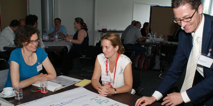 Teilnehmer sitzen am Gruppenarbeitstisch. Ein Teilnehmer steht am Tisch und legt den Stift aus der Hand, mit dem er die Diskussionsergebnisse der Diskutanten am Arbeitstisch auf das Posterpapier geschrieben hat. Die Teilnehmer schauen freundlich und lachen.