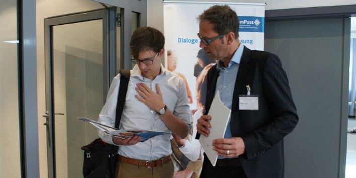 Zwei Teilnehmer stehen zusammen und schauen vertieft in die Veranstaltungsbroschüre. Im Hintergrund ist das Veranstaltungs Rollup sowie eine geöffnete Glastür zum Flur hin zu sehen.