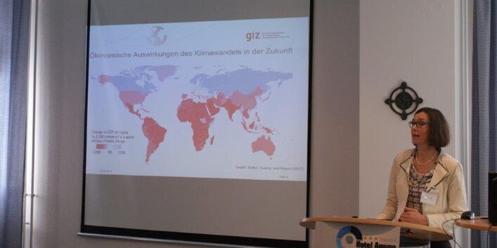 Vortragende steht am Rednerpult. Im Hintergrund ist eine Präsentationsbild in Form einer Weltkarte zu sehen.