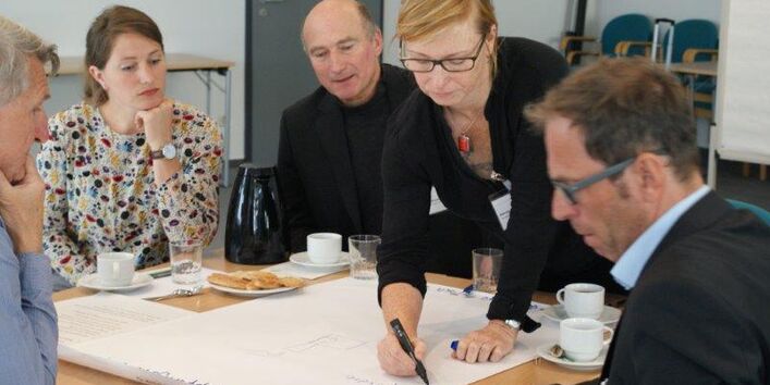 An einem Arbeitsgruppentisch steht eine Teilnehmerin und schreiben mit einem schwarzen Stift einen Beitrag auf Papier. Die anderen Teilnehmer schauen interessiertzu.