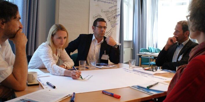 Teilnehmer sitzen an einem Gruppenarbeitstisch. Eine Teilnehmerin schreibt die ausgesprochenen Gedanken eines anderen Teilnehmers auf Papier. Andere Teilnehmer folgen dem interessiert.