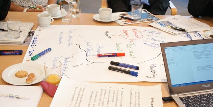 Ansicht auf einen Gruppenarbeitstisch zeigt in bunten Farben beschriebenes Papier, Stifte, ein aufgeklapptes Laptop, einen Teller mit Keksen und Gläser und Tassen mit Getränken.