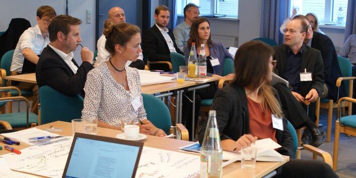 Der Blick geht hinein in den Arbeitsgruppenraum, wo die Teilnehmer an den Arbeitsgruppentischen sitzen. Ein Teilnehmer erzählt etwas. Andere Teilnehmer hören zu. Eine Teilnehmerin dreht sich dem Erzählenden zu.