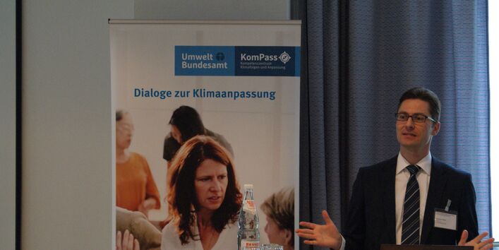 Vortragender steht mit seinen Händen gestikulierend vor einem Rollup Dialoge zur Klimaanpassung