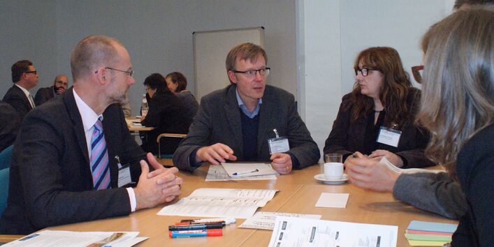 Teilnehmende sitzen am Gruppenarbeitstisch und sprechen miteinander. Auf dem Tisch befinden sich Moderationsmaterialien sowie Getränke.