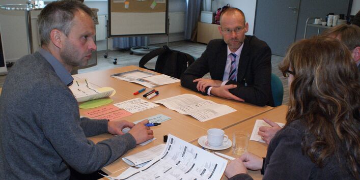 Teilnehmende sitzen am Gruppenarbeitstisch und schauen eine Teilnehmerin an. Moderationsmaterialien befinden sich auf dem Tisch sowie eine Kaffetasse und ein Getränkeglas.