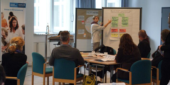 Teilnehmende trägt mithilfe einer Moderationswand vor. Sie zeigt auf die farbigen Karten und erläutert deren Bedeutung. Die anderen Teilnehmenden sitzen davor und hören zu.