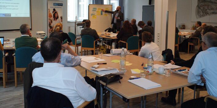 Im Vordergrund sitzen Teilnehmende an Gruppenarbeitstischen und hören dem Vortragenden zu. Der Vortragende steht im Hintergrund vo reiner Moderationswand und zeigt erläuternd auf die beschrifteten farbigen Moderationskarten.