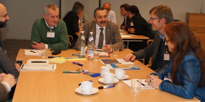 Teilnehmende sitzen am Gruppenarbeitstisch und schauen sich gegenseitig freundlich und lachend an. Auf dem Tisch stehen Tassen und Getränkegläser. Moderationsmaterialien wie farbige Stifte, Karten liegen auf dem Tisch.