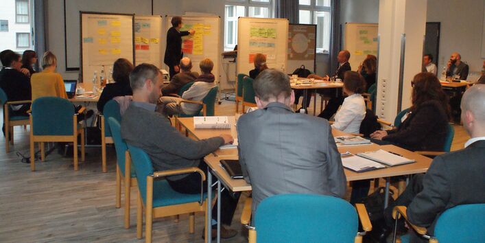Im Vordergrund sitzen die Teilnehmenden auf verschiedene Arbeitsgruppentischen verteilt im Raum und hören vertieft dem Vortragenden zu. Dieser steht vor einigen Moderationswänden, an denen beschriftete Moderationskarten hängen und zeigt erläuternd auf die Moderationswand. Der Blick geht im Hintergrund durch Fenster auf eine Häuserwand. 