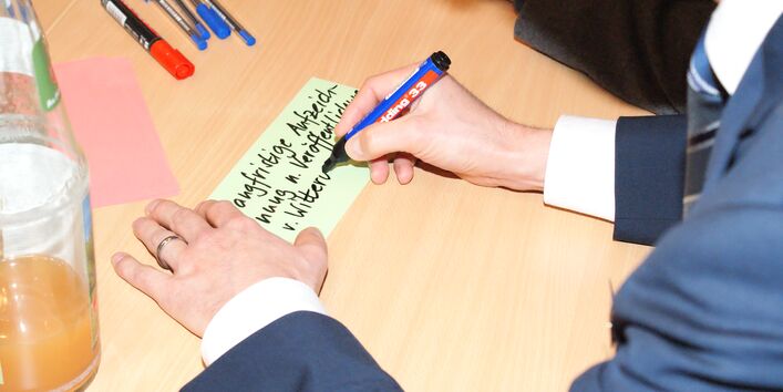 Teilnehmende beschriften farbige Moderationskarten in der Ansicht von oben sieht man die Hände. Farbige Moderationsstifte sowie eine Getränkeflasche sind ebenfalls zu sehen auf dem Gruppenarbeitstisch.