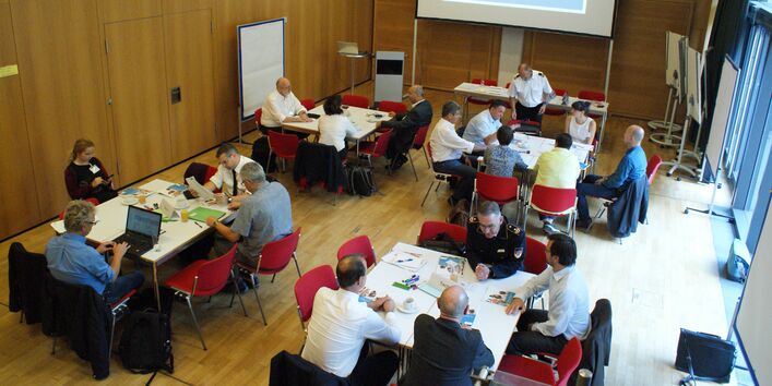 Die Aufnahme zeigt von oben die Teilnehmenden an Gruppenarbeitstischen sitzend in einem mit Holz vertäfelten Raum. Im Hintergrund ist das Veranstaltungsmotto Katastrophenschutz bei Trockenheit in der Landwirtschaft  in blau/weiss auf der Leinwand projiziert zu sehen.
