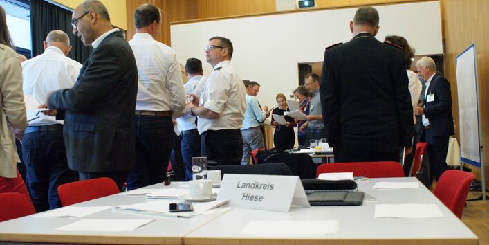 Teilnehemende stehen in lockeren Gesprächsgruppen beieinander. Sie tragen vorwiegend weiße Hemden und blaue Hosen. Im Vordergrund stehen ein Tischschild mit der Bezeichnung Landkreis Hiese.