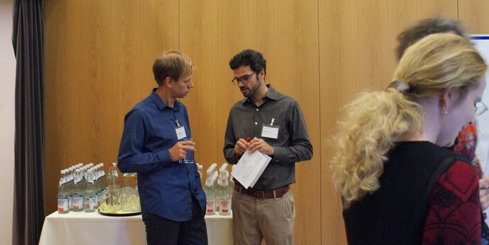 Zwei Teilnehmende stehen vor dem Getränketisch und unterhalten sich. Beide tragen Namenschilder am Hemd und halten Papiere und ein Glas Wasser in den Händen.