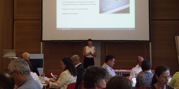 Teilnehmerin spricht vor der Präsentationsleinwand zu den anderen Teilnehmenden im verdunkelten Raum. Die Teilnehmenden sitzen an Gruppenarbeitstischen und schauen nachdenklich auf ihre Papiere.