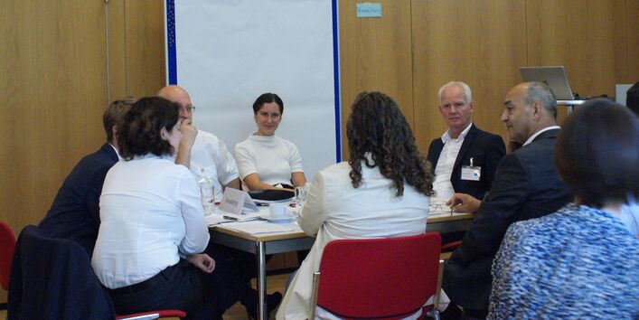 Teilnehmende sitzen an einem Gruppenarbeitstisch und hören einer Teilnehmerin zu. Im Hintergrund steht eine Pinwand mit einem großen leeren weißen Blatt bespannt.