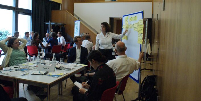 Im Vordergrund steht Gruppenarbeitstisch 3. Die Teilnehmenden sitzen um den Tisch herum und folgen den Ausführungen einer Teilnehmerin, die an der Präsentationswand zeigend steht. Im Hintergrund befindet sich eine große Fensterfront.