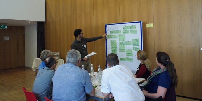 Ein Teilnehmer zeigt auf eine Präsentationswand mit grünen Moderationskarten. Im Vordergrund sitzen Teilnehmende am Gruppenarbeitstisch und hören zu.