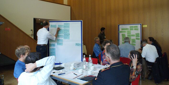 Ein Teilnehmender schreibt etwas auf eine Präsentationtswand, die mit blauen Moderationskarten bestückt ist. Im Raum sitzen alle Teilnehmenden an ihren Gruppenarbeitstischen.