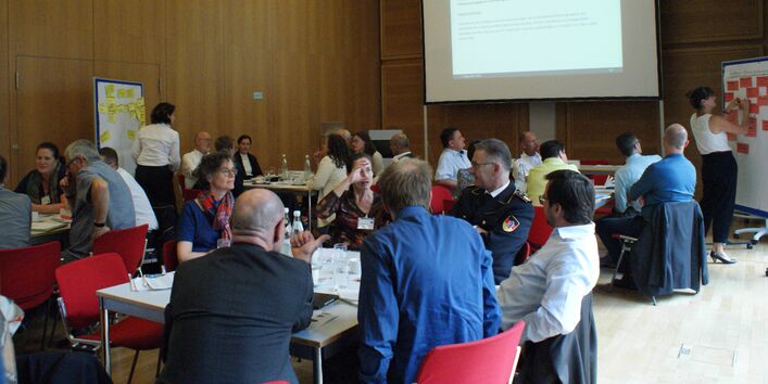 Der Veranstaltungsraum im Überblick zeigt alle Teilnehmenden an ihren Gruppenarbeitstischen und schauen zu, wie eine Teilnehmerin an einer Präsentationswand mit roten Moderationskarten präsentiert und etwas anschreibt. Auf der gegenüber liegenden Seite steht eine Präsentationswand mit gelben Moderationskarten.