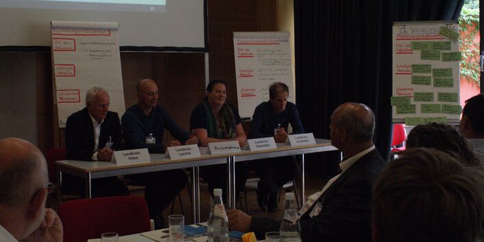 Auf einem Podium sitzen die teilnehmenden Vertreter der  Landkreise aufgereiht an einer Tischreihe. Namensschilder und Getränke stehen auf dem Tisch. Im Hintergrund stehen verschiedene Präsentationswände. Der Veranstaltungsraum ist präsentationsbedingt verdunkelt.