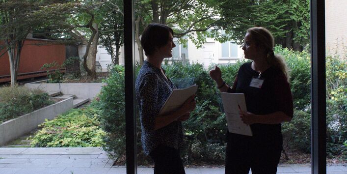 Zwei Teilnehmende stehen vor der Fensterfront und tauschen sich miteinander aus. Sie halten Unterlagen in den Händen sowie Stifte. Die Fensterfront gibt einen Blick frei ins Grüne.