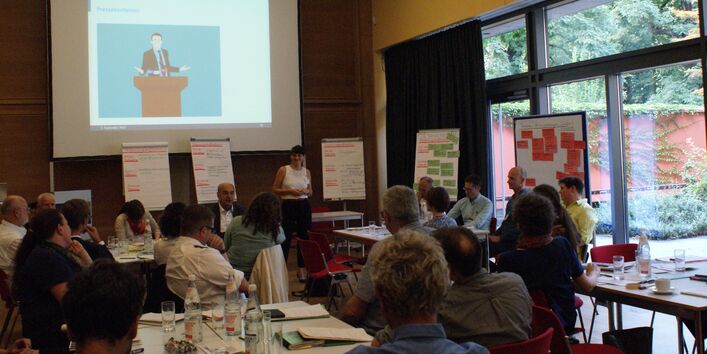 Die Teilnehmenden sitzen in lockerer Atmosphäre an ihren Arbeitstischen und hören einer Teilnehmerin zu, die am Kopfende des Raumes steht vor den aufgereihten Moderationswänden, die mit den farbigen Moderationskarten bestückt und beschriftet sind. Die große Fensterfront rechts davon gibt den Blick frei ins Grüne.