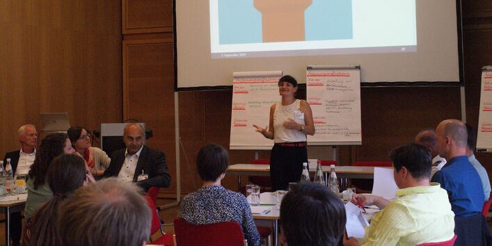Teilnehmerin spricht zu allen anderen Teilnehmenden. Sie steht gestikulierend vor beschrifteten Präsentationswänden. Im Vordergrund sitzen die Teilnehmenden und hören zu.