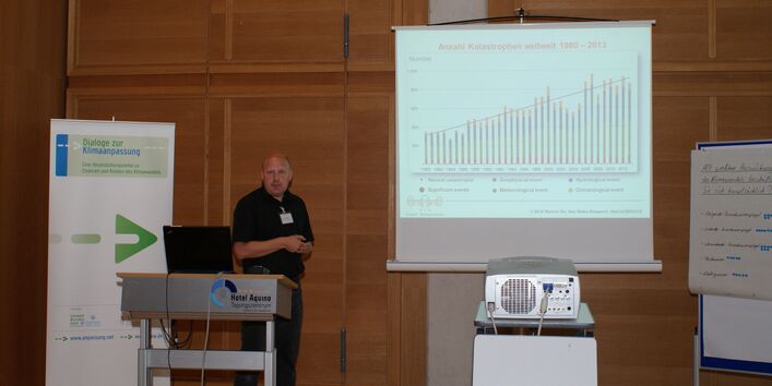 Vortragender steht am Rednerpult. Im Hintergrund stehen Moderationswände, Veranstaltungsaufsteller Dialoge zur Klimaanpassung. Die Präsentation sieht man im Hintergrund auf der Leinwand. Auf dem Rednerpult ist der geöffnete Deckel des Laptops sichtbar.