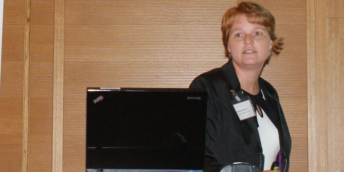 Vortragende steht vor holzvertäfelten Wand am Rednerpult. Das Rednerpult trägt vorne den Schriftzug Hotel Aquino Tagungszentrum.